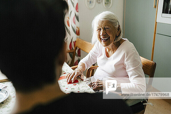 Fröhliche ältere Frau im Gespräch mit männlichem Pfleger beim Kartenspielen in der Küche