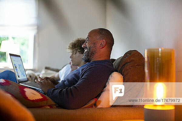 Smiling mature man using laptop while sitting by son on sofa in living room