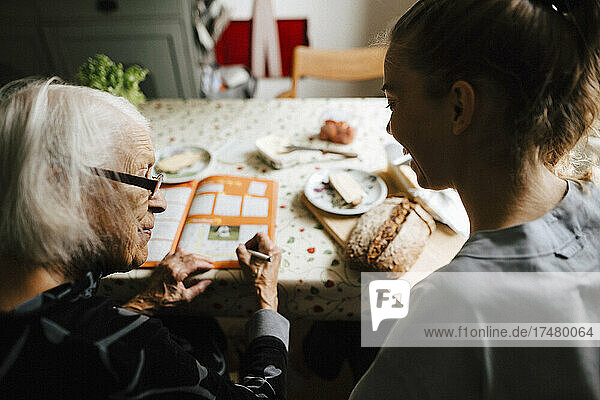 Elderly woman talking with female caregiver while solving crossword puzzle in kitchen