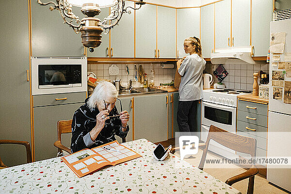 Ältere Frau mit Brille am Tisch  während eine Krankenschwester in der Küche steht