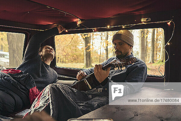 Mann spielt Gitarre  während sein Freund die Lichter im Campingbus betrachtet