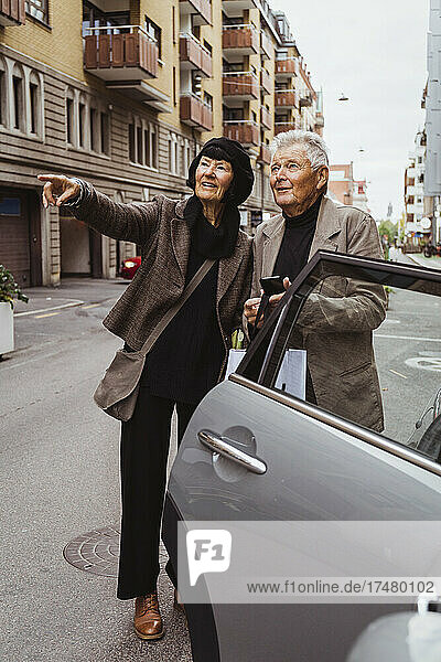 Lächelnde Frau  die auf ein Auto auf der Straße zeigt  während ein älterer Mann sein Smartphone in der Hand hält