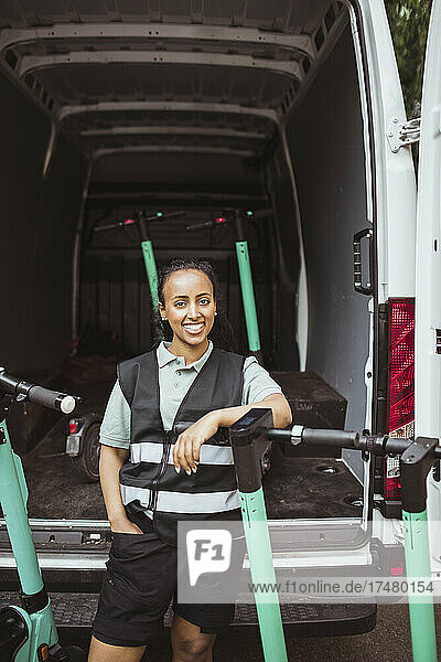 Lächelnde Zustellerin mit Hand in der Tasche  die sich auf einem Elektroroller gegen einen Lieferwagen lehnt