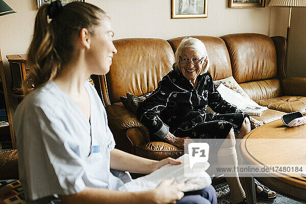 Lächelnde ältere Frau im Gespräch mit weiblicher Gesundheitspflege  während sie im Wohnzimmer zu Hause sitzt