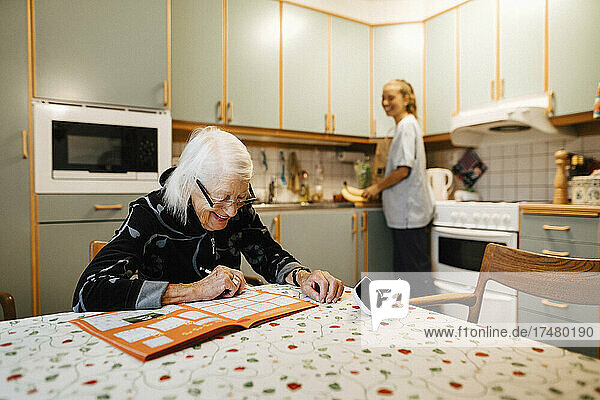 Lächelnde ältere Frau  die ein Sudoku in einem Buch löst  während eine weibliche Betreuerin zu Hause Küchenarbeiten erledigt