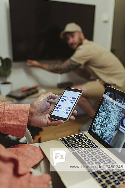 Älterer Mann benutzt ein Smartphone  während ein männlicher Arbeiter zu Hause den Fernseher repariert