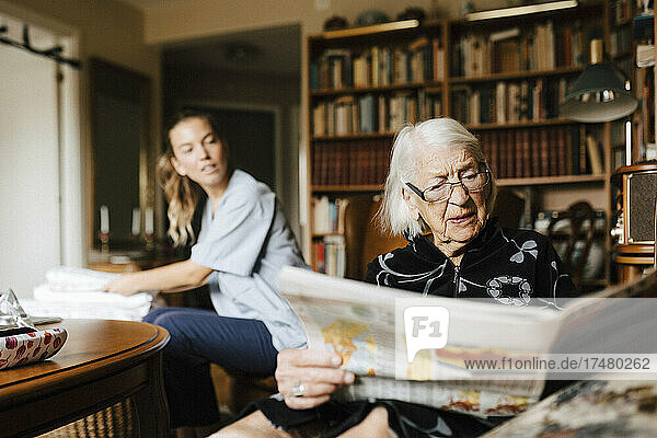 Senior woman reading newspaper while female caregiver looking at her in living room