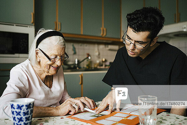 Young male healthcare worker playing crossword puzzle with senior woman in kitchen