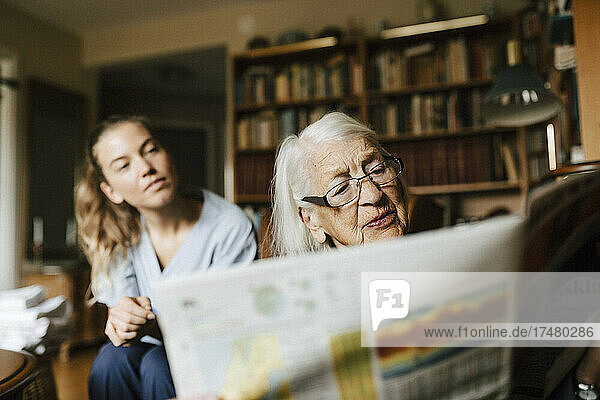 Ältere Frau liest Zeitung  die von einer Mitarbeiterin des Gesundheitswesens im Wohnzimmer gelesen wird