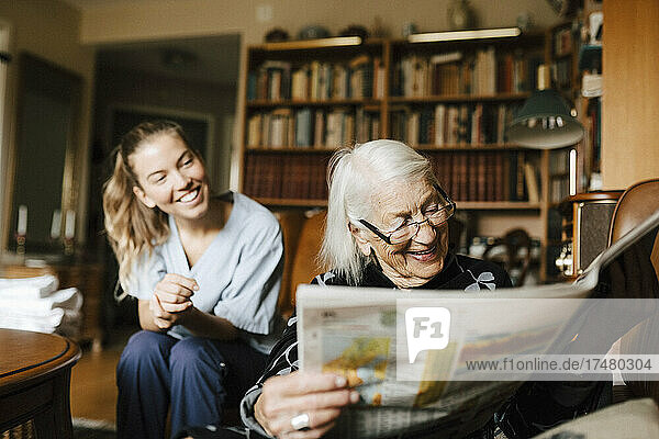 Lächelnde ältere Frau  die Zeitung liest  während im Hintergrund eine glückliche Betreuerin zu Hause ist