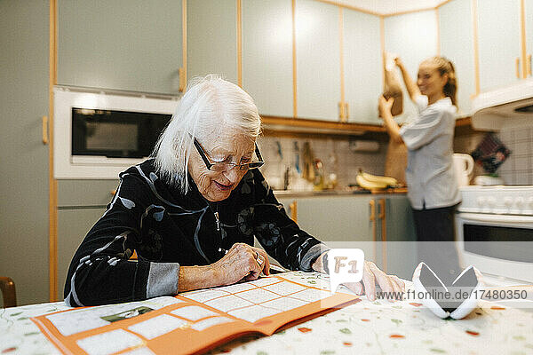 Eine ältere Frau löst ein Sudoku  während eine Krankenschwester in der Küche arbeitet