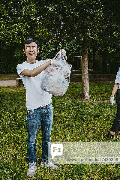 Ganzkörperporträt eines lächelnden jungen männlichen Aktivisten  der im Park eine Plastiktüte hält