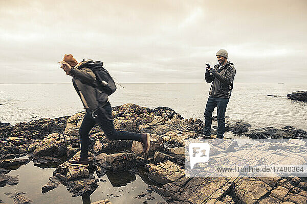 Man photographing male friend running on rocks by sea during sunset