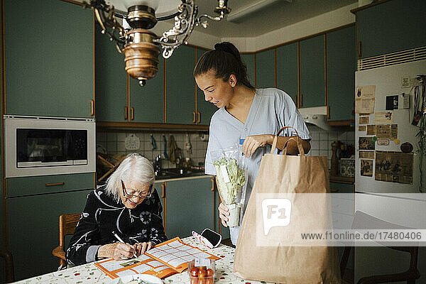 Junge Krankenschwester schaut  während eine ältere Frau zu Hause ein Kreuzworträtsel in einem Buch löst