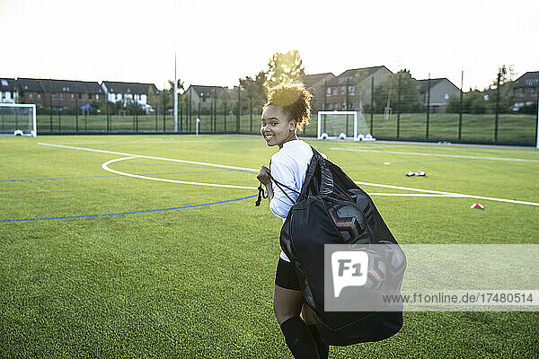 UK  Smiling female soccer player (12-13) carrying bag with balls in field