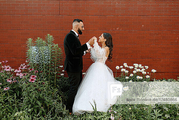Bride and groom holding hands against brick wall and flowers
