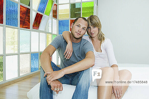 Mann und Frau sitzen umarmend auf einem Futonbett vor bunten Glasfenstern.