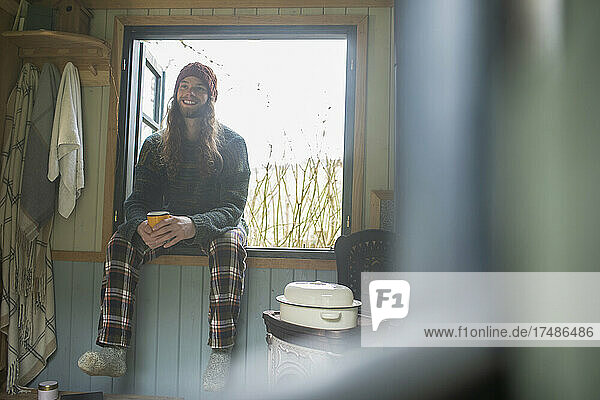 Fröhlicher junger Mann im Schlafanzug genießt Kaffee am Kabinenfenster