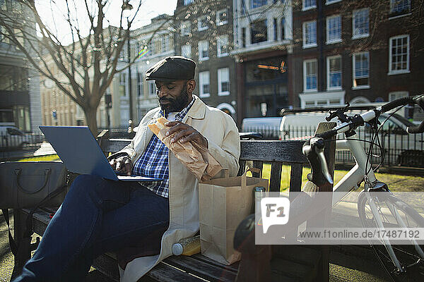 Businessman eating lunch and working at laptop in sunny city park