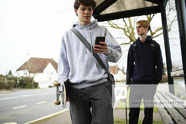 Teenager mit Skateboard und Smartphone an der Bushaltestelle