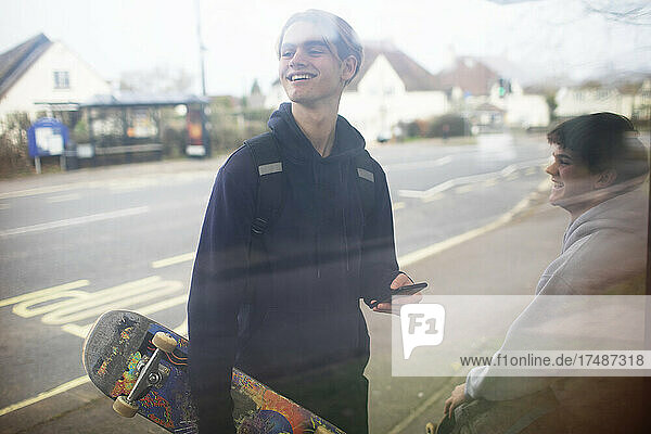 Glücklicher Jugendlicher mit Smartphone und Skateboard an der Bushaltestelle