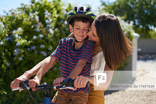 Glückliche  liebevolle Mutter  die ihren Sohn auf dem Fahrrad in einer sonnigen Einfahrt umarmt
