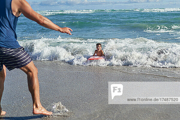 Vater beobachtet glücklichen Sohn beim Bodyboarden in der sonnigen Meeresbrandung