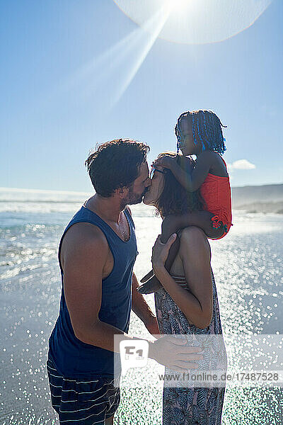 Zärtliches Paar mit Tochter küsst sich am sonnigen Meeresstrand