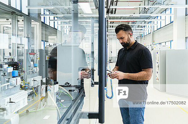 Engineer using mobile phone at industrial machine behind glass