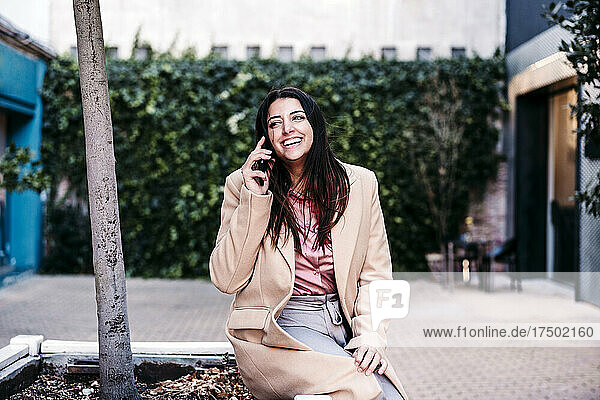 Smiling businesswoman talking on mobile phone by tree