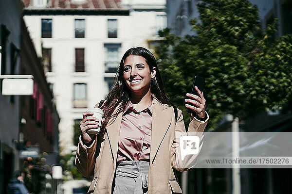 Young businesswoman with coffee cup and mobile phone on sunny day