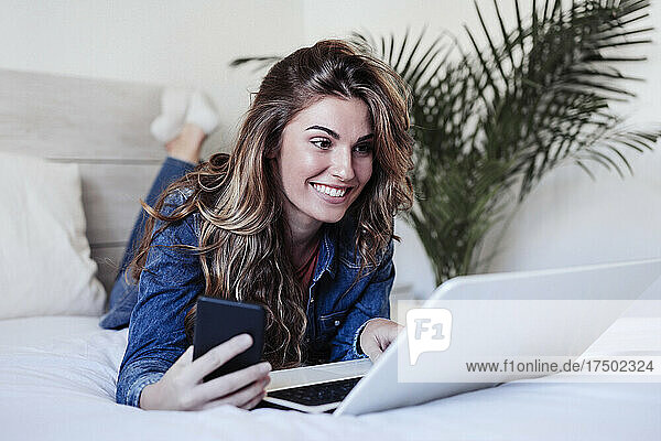 Lächelnde junge Frau mit langen Haaren benutzt Laptop im Bett