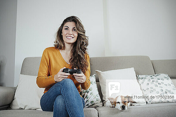 Beautiful woman playing video game sitting on sofa with dog at home