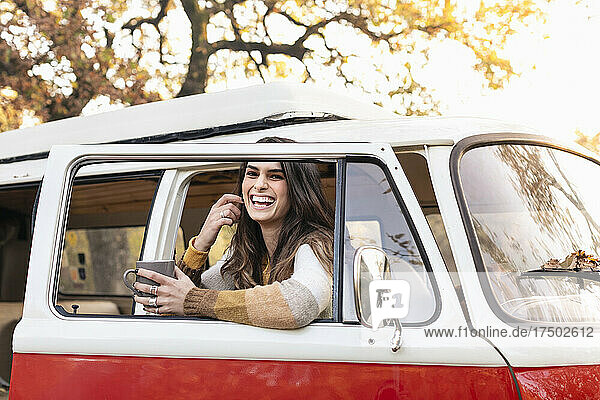 Cheerful woman with coffee mug leaning on door of van in autumn forest