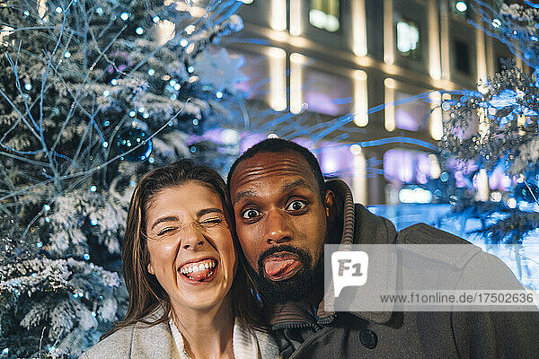 Playful couple sticking out tongues near Christmas tree at night
