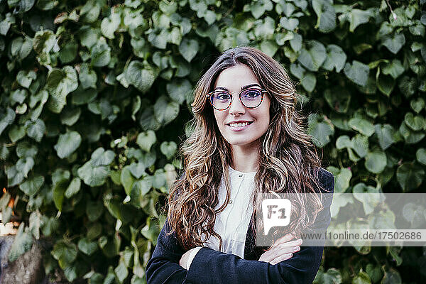 Young businesswoman with arms crossed in front of hedge