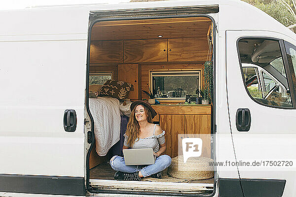 Lächelnde Frau sitzt mit gekreuzten Beinen und Laptop an der Tür des Wohnmobils