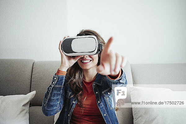 Smiling young woman wearing virtual reality headset and gesturing on sofa