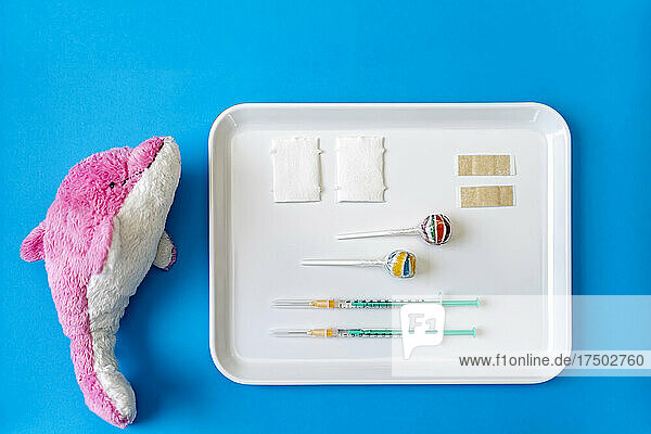 Stuffed dolphin toy by tray of vaccine injections and lollipop on blue background