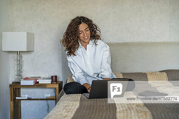 Smiling woman using laptop on bed in bedroom at home