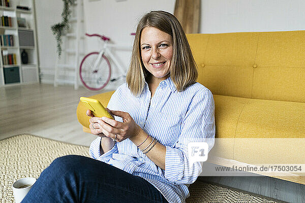 Smiling blond woman using mobile phone leaning on sofa at home