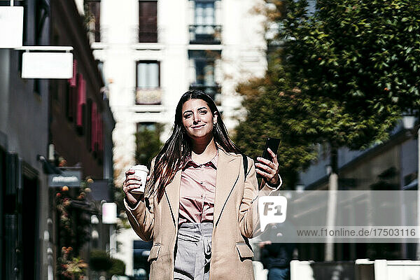 Smiling businesswoman holding disposable cup and mobile phone at city street