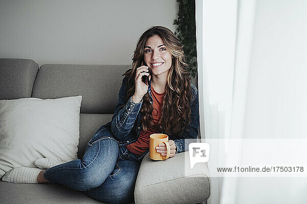 Smiling woman with coffee mug talking on smart phone at home