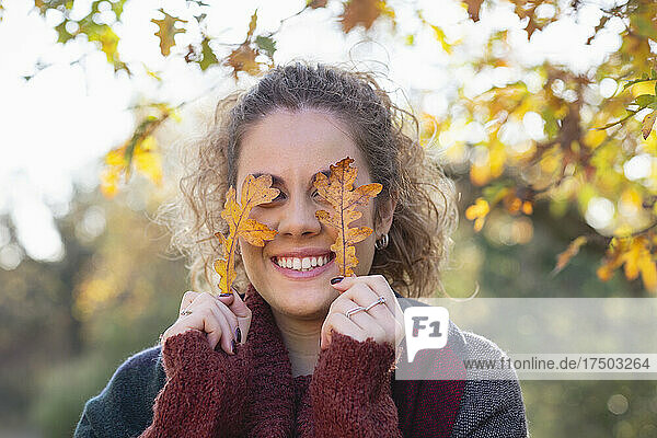 Smiling young woman holding autumn leaves in front of eyes at forest