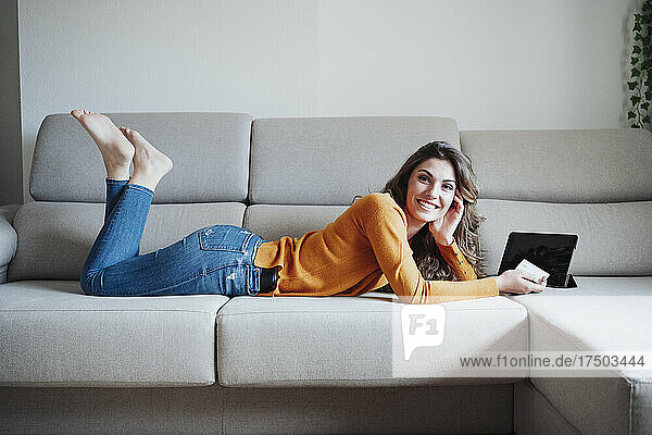 Young woman with credit card and tablet PC lying on sofa at home