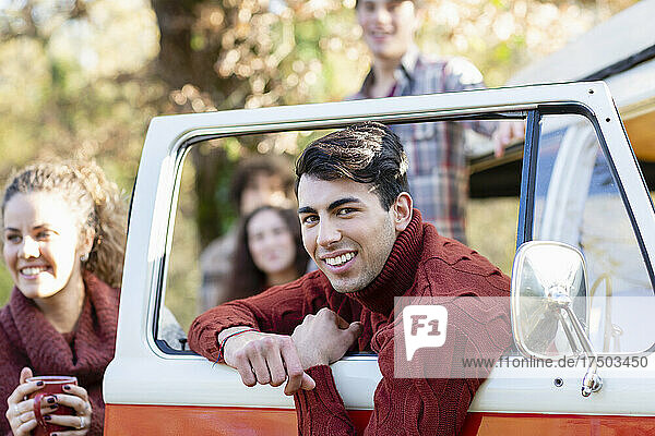 Smiling young man leaning on van's door with friends enjoying picnic in autumn forest
