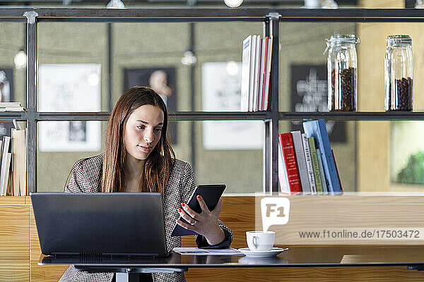 Young businesswoman with laptop using smart phone in cafe