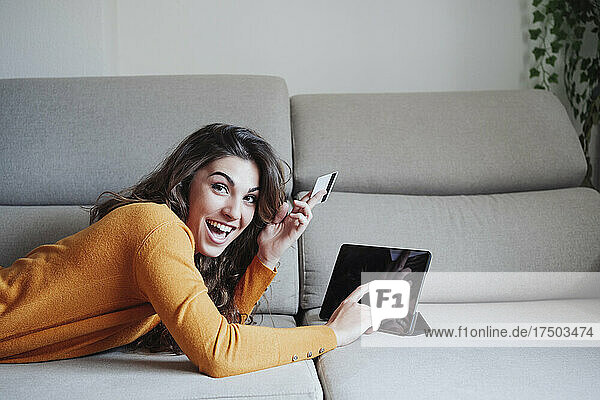 Excited woman using tablet PC holding credit card on sofa at home