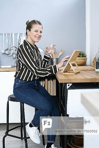 Lächelnde Frau sitzt auf einem Hocker mit einem Glas Wasser und nutzt ein digitales Tablet in der Küche