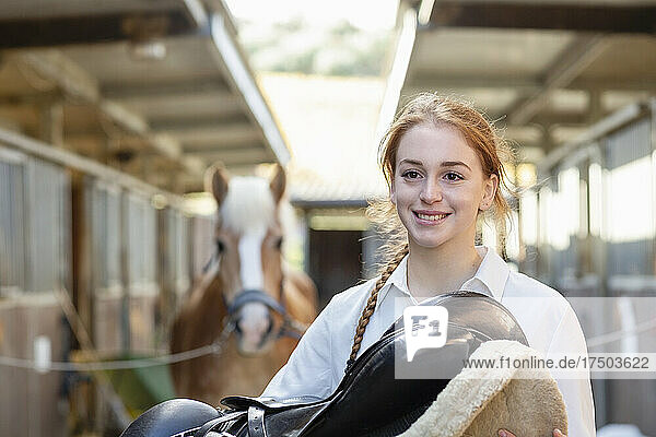Smiling young woman with saddle in stable